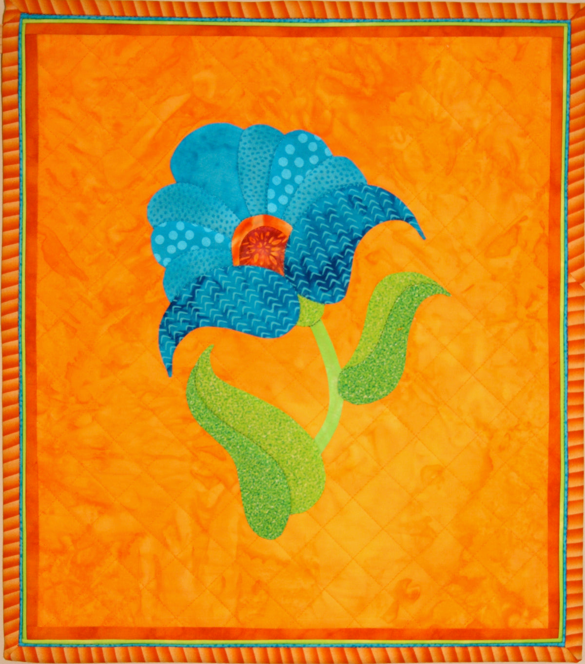 A square quilted fabric art piece featuring a stylized blue flower with green leaves on a bright orange background, finished with a decorative striped border.